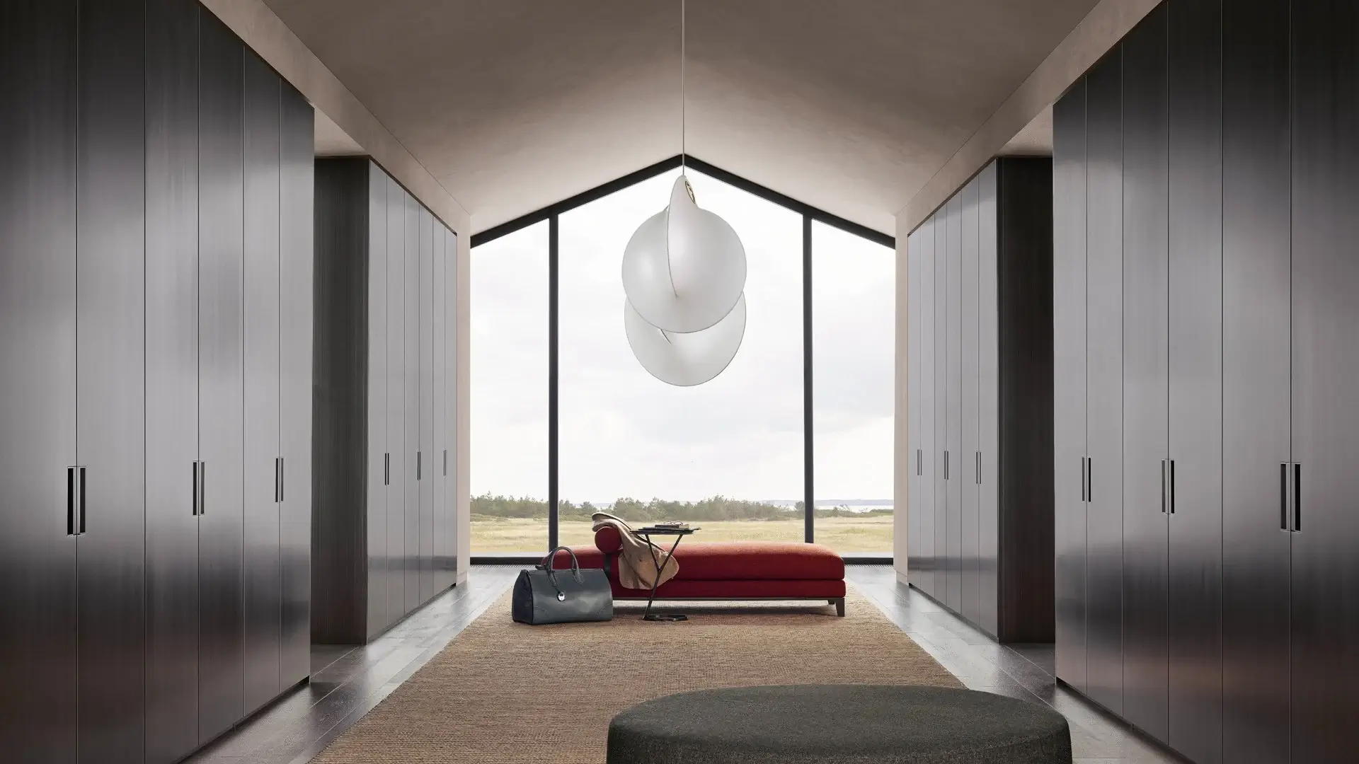 Luxury floor-to-ceiling dark wood wardrobe designs in a modern minimalist bedroom with a large window view, pendant lighting, and a red chaise lounge.