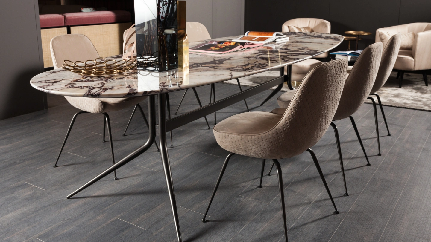 "A luxurious oval-shaped marble dining table with grey and purple veining, paired with six beige quilted upholstered chairs on sleek black metal legs, set in a modern dark-toned dining room by Ventura Interiors.