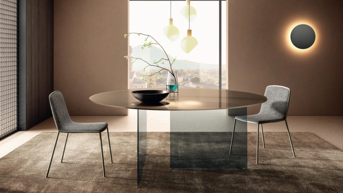 Modern glass dining table design with a tinted top and an architectural smoky base, surrounded by designer dining chairs and styled with a central vase, emphasizing the glass surface’s ability to highlight textures and maintain visual openness in small spaces.